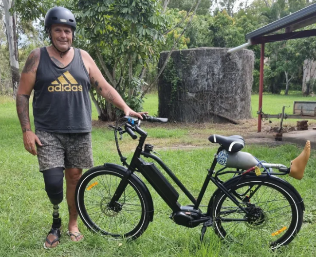 Man standing next to electric bike with one below knee prosthetic leg and another leg sitting on the rear rack of the bike