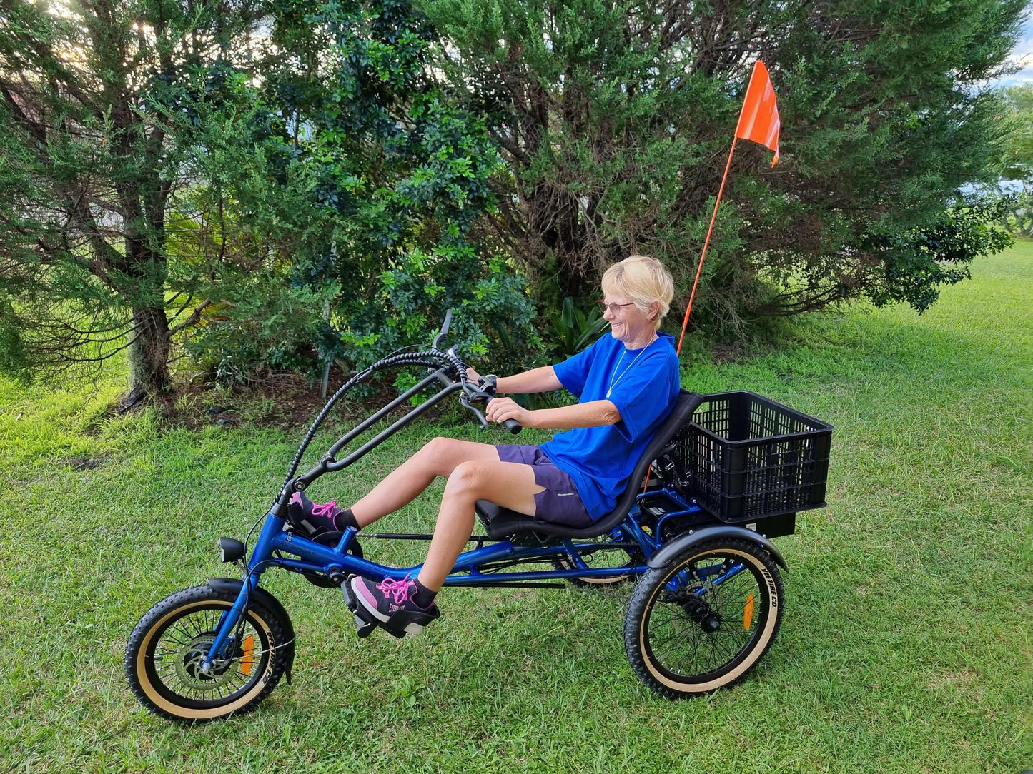 Women sitting on blue sit-down trike, semi-recumbent electric tricycle Trident with black crate at rear and orange flag