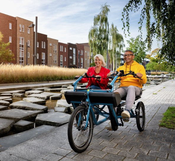 2 people smiling and having fun riding a huka orthros side-by-side trike. woman wearing red top, man wearing yellow top on a blue bike along a canal