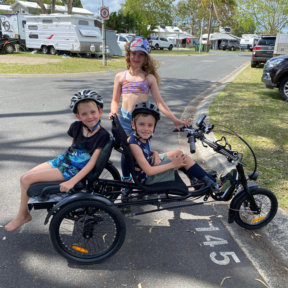 Three kids enjoying riding a trike in a caravan park, with one child with a disability riding the trike, one sitting on the back and another standing next to the trike