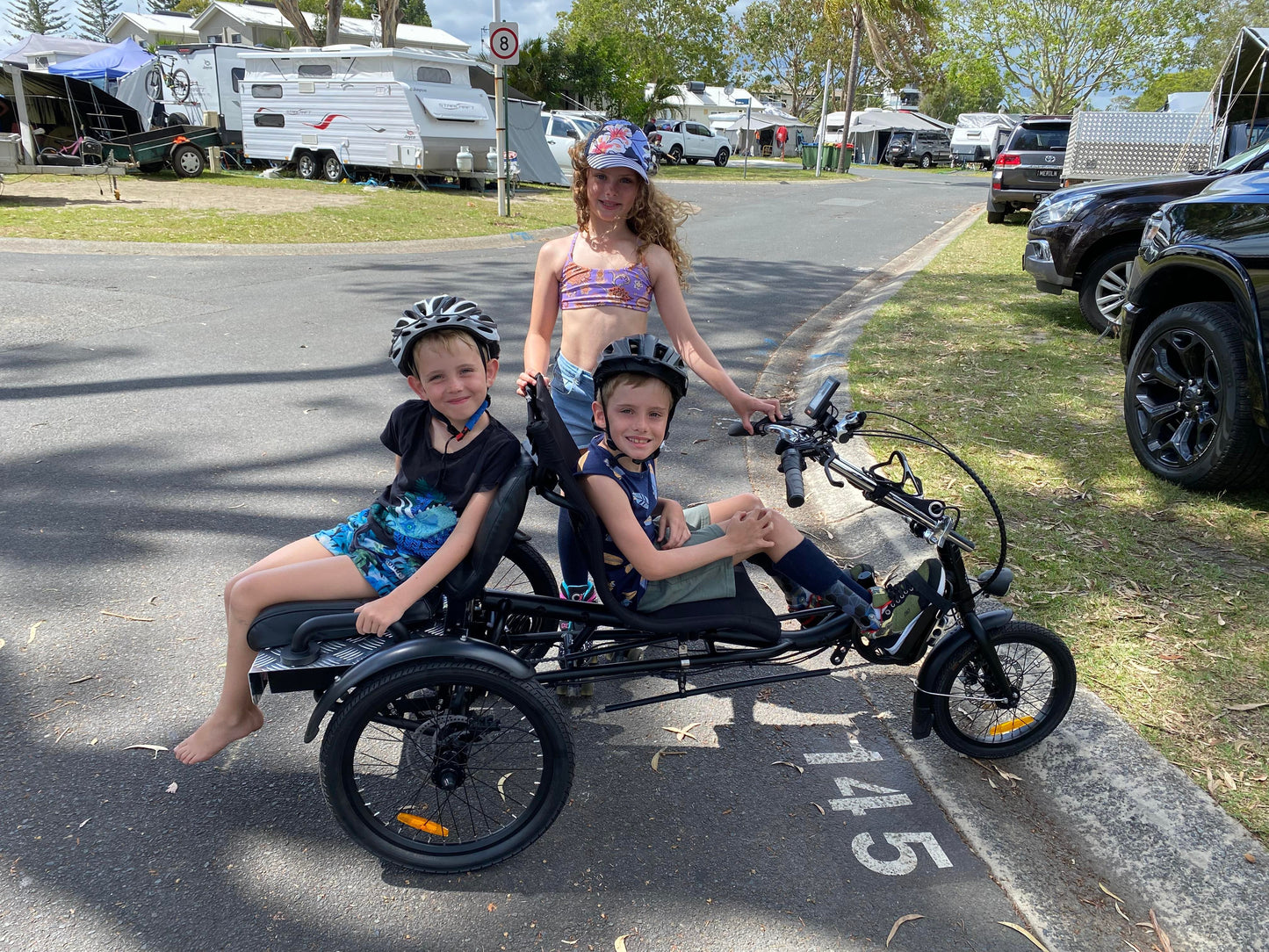 Three kids enjoying riding a trike in a caravan park, with one child with a disability riding the trike, one sitting on the back and another standing next to the trike