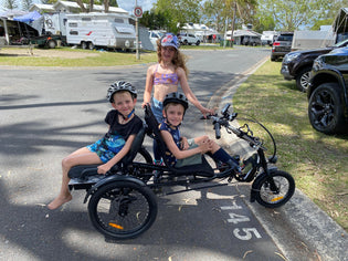  Three kids enjoying riding a trike in a caravan park, with one child with a disability riding the trike, one sitting on the back and another standing next to the trike
