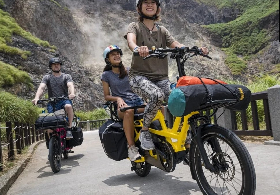 Electric cargo bike carrying woman and small girl and lots of luggage and another rider on another bike also carrying cargo