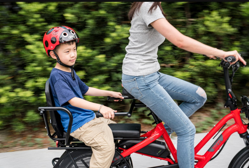 Small boy wearing blue tshirt and red helmet riding behind a woman on a red electric cargo bike