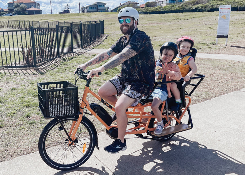 
                      
                        Happy family with dad riding a orange Tribe Bikes Evamos Longtail Cargo Bike with two smiling happy kids
                      
                    